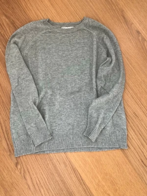 Grå stickad tröja i ullmix - Mysig grå stickad tröja med rund hals och långa ärmar. Tröjan har ribbstickade muddar vid ärmslut och nederkant. Perfekt för lager på lager och enkel att matcha till olika outfits.