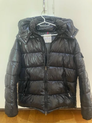 Svart dunjacka från Moncler - Snygg svart dunjacka från Moncler med glansig finish, huva och dragkedja framtill. Jackan har två fickor med dragkedja och klassisk Moncler-logga på ärmen. Perfekt för kalla vinterdagar och riktigt trendig stil.
