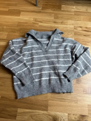 Grå randig stickad tröja med krage - Mysig grå stickad tröja med vita horisontella ränder och bred krage. Tröjan har v-ringning, långa ärmar och ribbstickade muddar vid ärmslut och nederkant. 
