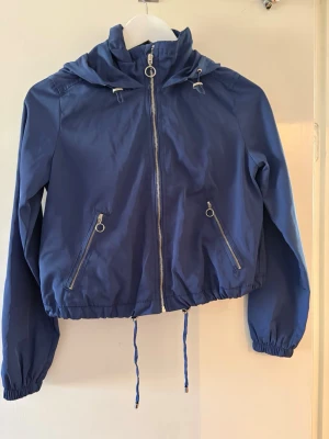 Blå jacka med huva och dragkedja - Snygg blå jacka med huva och dragsko i midjan. Jackan har två fickor med dragkedja och coola ringdetaljer på både fickor och dragkedja. Materialet är tunt och lätt, perfekt för lager på lager. Elastiska muddar vid ärmslut ger en skön passform.