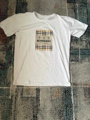 Vit Burberry t-shirt med rutigt tryck - Snygg vit t-shirt från Burberry med det klassiska rutiga mönstret och logga på bröstet. T-shirten har korta ärmar och rund halsringning. Perfekt för dig som gillar stilrena plagg med ikoniska detaljer.