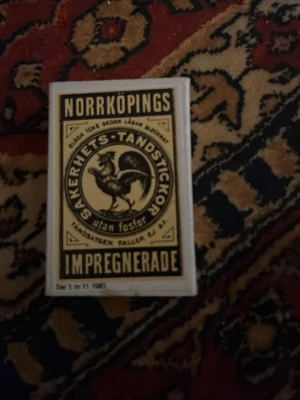 Norrköpings säkerhetständstickor ask - Unik tändsticksask med klassisk Norrköpings-etikett och illustration av en tupp. Framsidan har svart och gul design med texten 'Säkerhets-Tändstickor'. Baksidan listar gamla etiketter och motiv från olika år. Perfekt för samlare av retroprylar.
