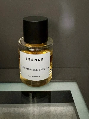 ESSNCE Irresistible Enigma parfym - En stilren parfymflaska från ESSNCE med doften Irresistible Enigma. Flaskan är cylindrisk i genomskinligt glas med svart lock och vit etikett. Innehållet är gyllene och flaskan har en modern, minimalistisk design. Eau de Parfum. Förvarad mörkt och svalt