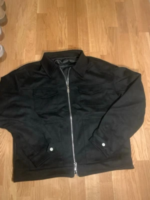 Svart med dragkedja - Stilren svart jeansjacka med klassisk krage, två bröstfickor med lock och silverfärgad dragkedja framtill. Jackan har knappar vid ärmsluten och är helfodrad för extra komfort. Perfekt för dig som gillar en clean och tidlös look.
