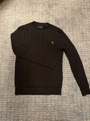 Svart kabelstickad tröja GANT - Snygg svart kabelstickad tröja från GANT med rund halsringning och broderad logga på bröstet. Tröjan har ribbade muddar och är tillverkad i ett mjukt stickat material. Den är otroligt snygg och i väldigt fint skick, nästan aldrig använd. Skriv vid funderingar eller frågor.