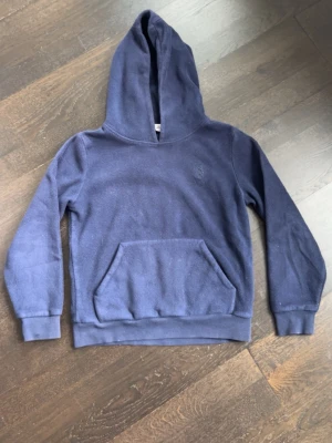 Mörkblå fleece hoodie med magficka - Mysig mörkblå hoodie i fleece med stor huva och klassisk magficka. Perfekt för chill dagar eller när du vill hålla dig varm. Diskret broderad logga på bröstet och ribbade muddar vid ärmslut och nederkant. 