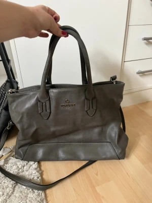 Grå handväska vintage  - Snygg grå handväska från Vagabond i slätt konstläder. Väskan har både handtag och en avtagbar axelrem, samt dragkedja upptill. Perfekt för dig som vill ha en stilren och rymlig väska med klassisk design och diskret logga framtill.