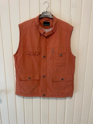 Orange väst från Frank Eden - Snygg orange väst från Frank Eden med flera praktiska fickor framtill och hög krage. Perfekt för dig som gillar funktionella detaljer och en avslappnad stil. Västen har knapp- och dragkedjestängning samt meshfoder på insidan.