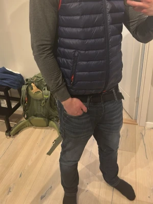 Mörkblå dunväst med dragkedja från Ralph Lauren - Snygg mörkblå dunväst med quiltad design och dragkedja framtill. Västen har två fickor med dragkedja och röda detaljer vid fickorna. Perfekt att slänga över en hoodie eller långärmad tröja för extra värme.