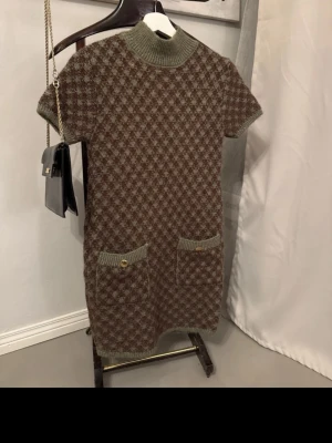 stickad kortklänning - Kort stickad klänning i brunt och grönt med rutigt mönster, ribbad polokrage och korta ärmar. Två fickor framtill med dekorativa knappar. Perfekt för dig som gillar retrovibbar och mysiga material.