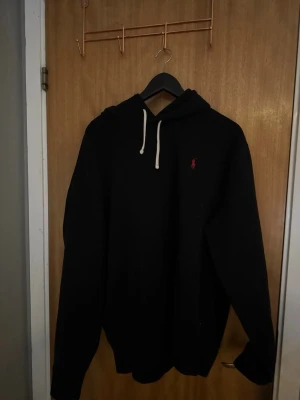 Svart hoodie från Polo Ralph Lauren - Svart hoodie från Polo Ralph Lauren med vit dragsko och klassisk röd logga broderad på bröstet. Mjuk bomullskänsla och avslappnad passform, perfekt för en chill stil.