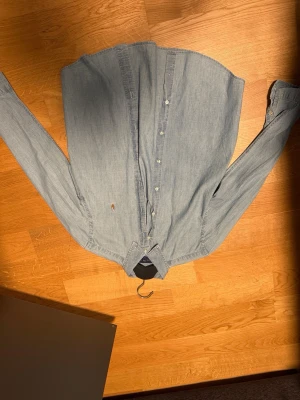 Ljusblå skjorta från Polo Ralph Lauren - Klassisk ljusblå skjorta från Polo Ralph Lauren med broderad logga på bröstet. Skjortan har knappar framtill, lång ärm och en avslappnad passform. Perfekt för en stilren och tidlös look.