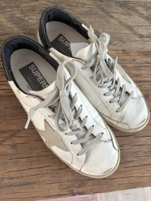 Golden Goose i modellen Superstar vita sneakers med stjärna - Snygga vita sneakers från Golden Goose med grå mockastjärna på sidan, grå skosnören och svart hälparti. Skorna har en klassisk låg modell och är tillverkade i läder med detaljer i mocka. Perfekta för en trendig och avslappnad stil.