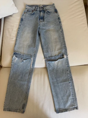 Ljusblå mom jeans med hål på knäna - Säljer ett par ljusblå mom jeans med hög midja och raka ben. Jeansen har stora slitna hål över båda knäna och klassiska fem fickor. Perfekt för en avslappnad och trendig look. Materialet är denim i bomull.