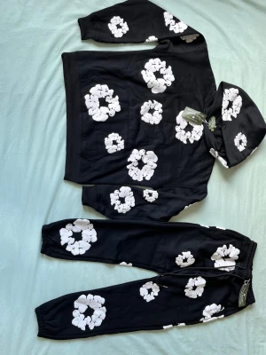 Svart loungewear-set med vita blommor - Snyggt svart loungewear-set från Daily Paper med vita blommönster över hela både hoodie och byxor. Hoodien har huva och magficka, byxorna har resår i midja och benslut. Perfekt för chill dagar hemma eller när du vill vara bekväm men ändå se cool ut.