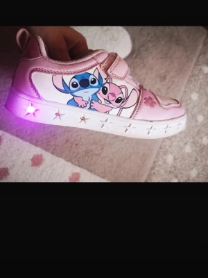 Rosa sneakers med Stitch-motiv och LED - Snygga rosa sneakers med motiv av Stitch och Angel från Disney. Skorna har vita sulor med stjärndetaljer och inbyggda LED-lampor som lyser i lila. Kardborreknäppning och syntetmaterial. Perfekta för dig som gillar färg och coola detaljer!