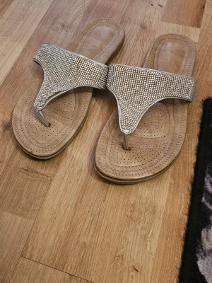 Beige sandaler med strassdetaljer - Snygga beige sandaler med breda remmar täckta av glittrande strass. Modellen är en klassisk flip-flop med mjuk, perforerad sula för extra komfort. Perfekta för dig som vill ha något som sticker ut och glänser lite extra. Materialet är syntet och textil.