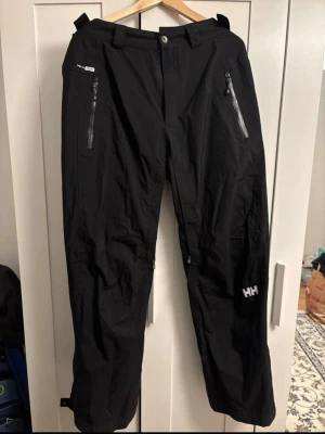 Helly Hansen svarta täckbyxor - Svarta täckbyxor från Helly Hansen med raka ben, två vattentäta dragkedjefickor framtill och justerbara benslut. Byxorna är gjorda i slitstarkt, vattenavvisande material och har HH-logga på benet. Perfekta för vinterns aktiviteter utomhus.