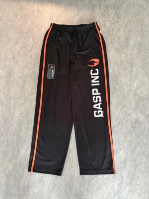 Svarta träningsbyxor från GASP INC - Säljer ett par svarta träningsbyxor från GASP INC med vita tryck på ena benet och orangea detaljer längs sidorna. Byxorna har dragsko i midjan och raka ben. Perfekta för gymmet eller annan träning.