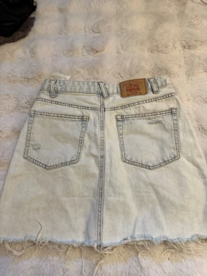 Ljusblå jeanskjol från Pull&Bear - Jeanskjol från Pull&Bear i klassisk denim med rå kant nertill och femficksmodell. Kjolen har knapp- och dragkedjestängning. Har en liten fläck på baksidan men utöver det så har den inga defekter