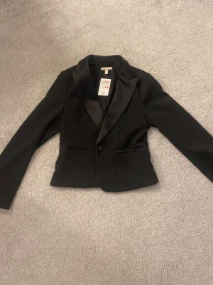 Svart kavaj från H&M, storlek 36 - Stilren svart kavaj från H&M i storlek 36. Klassisk modell med smala slag, enkelknäppning och två fickor framtill. Säljer då den aldrig kommit till användning😊
