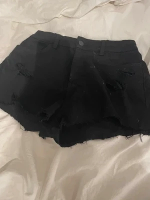 Svarta slitna shorts med rå kant - Säljer ett par svarta shorts med rå kant och slitna detaljer. De har bälteshällor och knappstängning framtill. Perfekta för en avslappnad och edgy stil. Materialet känns som bomull eller denim.