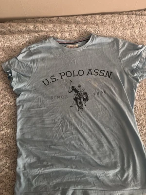 Ljusblå t-shirt från U.S.Polo Assn. Läs beskrivning  - Ljusblå t-shirt från U.S. Polo Assn. med klassiskt tryck framtill och texten 'Since 1890'. T-shirten har rund halsringning och korta ärmar. Tillverkad i mjuk bomull för en bekväm känsla. Den är jättefin och skön ha på sig size M använd men syns inte på det! Den har liten sträck klår tecken vit färg men syns inte alls alls förutom om man är väldig nära tröjan 