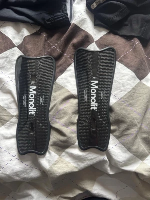 Monolit benskydd i kolfiber - Säljer ett par svarta benskydd från Monolit i kolfiber med vit logga. De är lätta och har en ergonomisk form som passar smidigt på benet. Perfekta för fotboll eller annan sport där du behöver skydda smalbenen. Snygg och modern design. 19cm