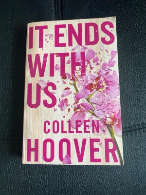 It Ends With Us av Colleen Hoover - En gripande roman av Colleen Hoover med omslag i träliknande beige och rosa toner, prydd med en krossad orkidé. Boken är på engelska och handlar om kärlek, svåra val och relationer. Perfekt för dig som gillar starka känslor och moderna berättelser.