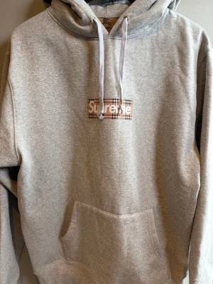 Grå Supreme x Burberry hoodie - Snygg grå hoodie från Supreme och Burberry med klassiskt rutigt Burberry-mönster bakom Supreme-loggan på bröstet. Hoodien har huva med dragsko och stor magficka. Perfekt för dig som gillar streetwear och exklusiva samarbeten.