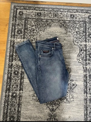 Blå skinny jeans från Tommy Hilfiger - Säljer ett par blå skinny jeans från Tommy Hilfiger med klassisk femficksdesign och diskret logga på bakfickan. Jeansen har en snygg tvättad look och smal passform som sitter tajt längs benen. Perfekta för en trendig och avslappnad stil.