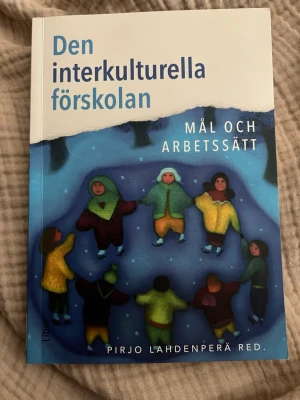Den interkulturella förskolan - Boken 'Den interkulturella förskolan: Mål och arbetssätt' av Pirjo Lahdenperä handlar om interkulturalitet i förskolan och pedagogiska metoder. Omslaget är blått och vitt med en illustration av barn som håller varandra i händerna. Mjukt pärmomslag.