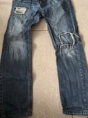 Blå slitna jeans med hål - Säljer ett par blå jeans med slitningar och stora hål på benen för en cool, avslappnad look. Jeansen har raka ben, dubbla bakfickor och detaljerade sömmar. Perfekta för dig som gillar streetstyle och vill ha ett par statement-jeans i garderoben.