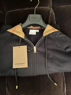 Svart hoodie från Burberry med dragkedja - Svart hoodie från Burberry med dragkedja, klassisk beige huva med Burberrys rutiga mönster på insidan. Guldiga detaljer på snören och dragkedja. Mjuk bomullskänsla och fickor framtill. Perfekt för en stilren och lyxig streetlook.