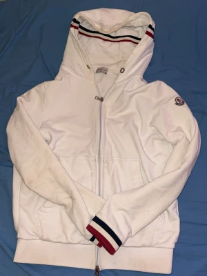 Vit hoodie från Moncler med dragkedja ÄKTA - Snygg vit hoodie från Moncler med hel dragkedja, huva och ribbade muddar med röd och marinblå rand. Klassisk Moncler-logga på ärmen och innerfoder i huvan med röd och blå rand. Perfekt för en stilren och sportig look.