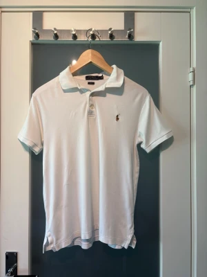 Vit pikétröja från Ralph Lauren - Klassisk vit pikétröja från Ralph Lauren med korta ärmar och krage. Tröjan har knappar framtill och den ikoniska broderade loggan på bröstet. Perfekt till jeans eller shorts för en clean och stilren look.
