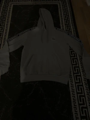 Grå EA7 hoodie med logga - Snygg grå hoodie från EA7 med huva och magficka. Svarta och vita EA7-band längs ärmarna och liten EA7-logga på bröstet. Perfekt för en chill och sportig stil. Mjuk bomullskänsla och ribbade muddar vid ärmslut och midja.