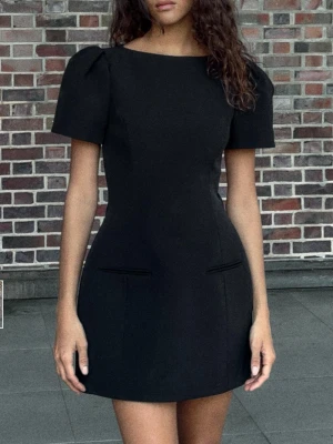 Svart klänning Zara - Stilren svart klänning från Zara med korta puffärmar och rund halsringning. Klänningen har en figurnära passform och slät design utan mönster, samt diskreta fickdetaljer framtill. Perfekt för dig som gillar minimalistisk och chic stil.