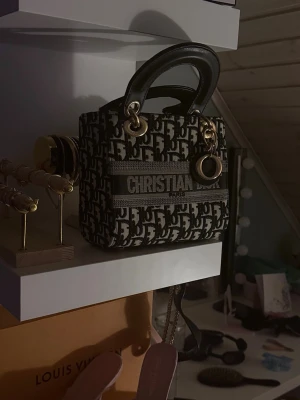 monogram handväska - Säljer en ikonisk handväska  med svart och beige monogrammönster, guldfärgade metalldetaljer och rejäla svarta handtag. Väskan har tydlig logga framtill och klassiska berlocker. Perfekt storlek för det viktigaste och riktigt snygg statement piece. Snabb köp så säljer jag den för 1150kr
