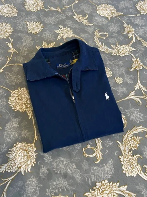 Marinblå jacka från Polo Ralph Lauren - Stilren marinblå jacka med dragkedja från Polo Ralph Lauren. Jackan har klassisk krage, rutigt innerfoder och den ikoniska vita logotypen broderad på bröstet. Perfekt för dig som gillar tidlös och snygg design.