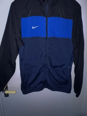 Nike träningsjacka blå och svart - Nike träningsjacka med dragkedja i svart och mörkblått, med en blå panel över bröstet och vit Nike-logga. Långärmad modell i lätt och sportigt material, perfekt för träning eller uppvärmning.