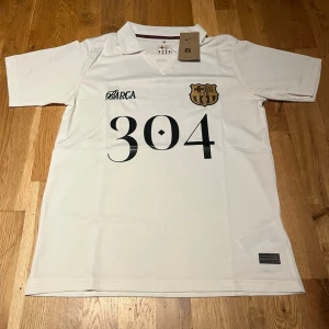 Barcelona X lamine yamal special edition  - Vit kortärmad FC Barcelona tröja från Nike med svart tryck '304' och klubbmärke på bröstet. På ryggen finns en stor illustration med texten 'Lamine Yamal'. Perfekt för dig som älskar fotboll och vill sticka ut på planen.