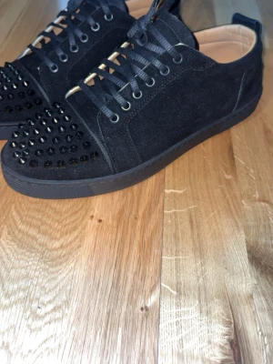 Svarta Louboutin sneakers med nitar - Säljer ett par svarta sneakers från Christian Louboutin i mocka med coola svarta nitar på tån. Skorna har svarta snören, låg profil och beige innersula med röd logga. Perfekta för dig som vill sticka ut med en edgy stil.