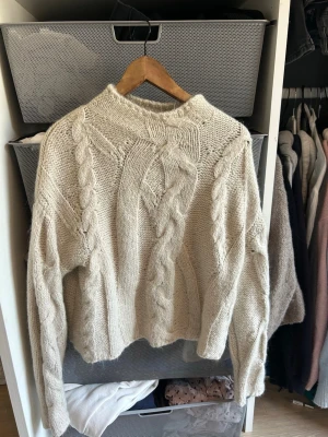 Beige kabelstickad tröja - Mysig beige stickad tröja med grova kabelstickade detaljer framtill och längs ärmarna. Tröjan har en något högre hals och är tillverkad i ett tjockt, mjukt garn som ger en avslappnad och trendig look. (Är man något känslig för saker på nacken/halsen och speciella garn rekommenderar jag kanske inte denna tröja). Perfekt för kyliga dagar eller sommarkvällar🤗☀️Pris kan absolut diskuteras!😊