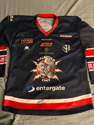 Halmstad Hammers hockeytröja Bauer - Säljer en mörkblå Halmstad Hammers hockeytröja från Bauer med röd krage, samt röda och vita detaljer på ärmar och nederkant. Tröjan har flera sponsormärken, stort klubbmärke på bröstet och tryck både fram och bak. Materialet är glansigt och typiskt för matchtröjor. Tröjan har även aotogtafer från några av spelarna i laget. 