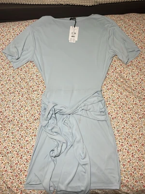 Ljusblå kortklänning från Gina Tricot - Säljer en ljusblå kortklänning från Gina Tricot med knyt i midjan och korta ärmar. Klänningen har en enkel och stilren design som passar perfekt för dig som gillar en clean look. Materialet är mjukt och följsamt, vilket gör den bekväm att bära.
