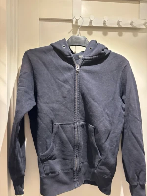Mörkblå hoodie med dragkedja Uniqlo | strl XS - Mörkblå hoodie från Uniqlo i storlek XS. Klassisk modell med dragkedja framtill, huva med snörning och två stora fickor. Tillverkad i mjukt bomullsmaterial som är skönt mot huden. Perfekt för en avslappnad och stilren look.