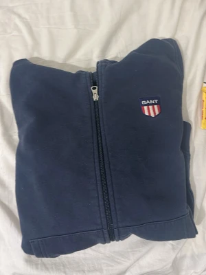 Mörkblå hoodie med dragkedja från GANT - Mörkblå hoodie från GANT med hel dragkedja och broderad logotyp på bröstet. Klassisk modell med huva och kängurufickor framtill. Mjuk bomullsmix som är skön att ha på sig, perfekt för en avslappnad stil.