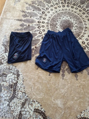 Psg shorts 2 för 50 - Säljer ett par mörkblå shorts från PSG med klubbens logga på vänster ben. Shortsen har elastisk midja och är tillverkade i ett lätt och svalt polyestermaterial, perfekta för sport eller chill. 140cm och den andra S