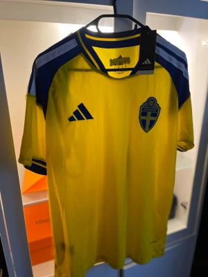 Sverige matchtröja Adidas gul/blå M - Säljer en officiell Sverige matchtröja från Adidas i storlek M. Tröjan är gul med blå detaljer, har korta ärmar och Sveriges landslagsemblem på bröstet. Tillverkad i ett lätt och ventilerande material, perfekt för fotboll eller träning.